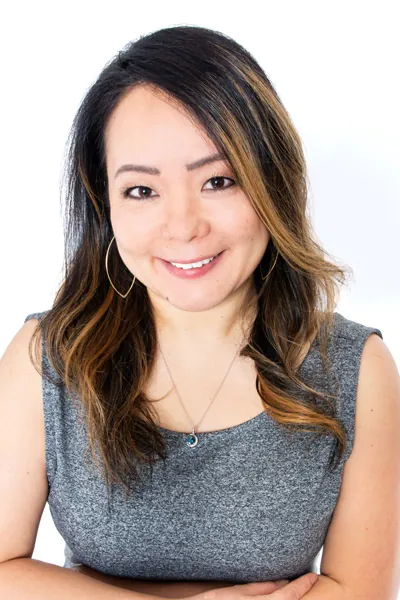 A smiling woman with long, wavy hair, wearing a grey top and necklace, poses against a plain white background.