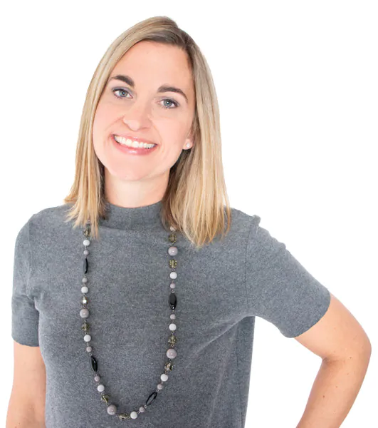 A woman with blonde hair smiles while wearing a gray top and a long beaded necklace against a white background.