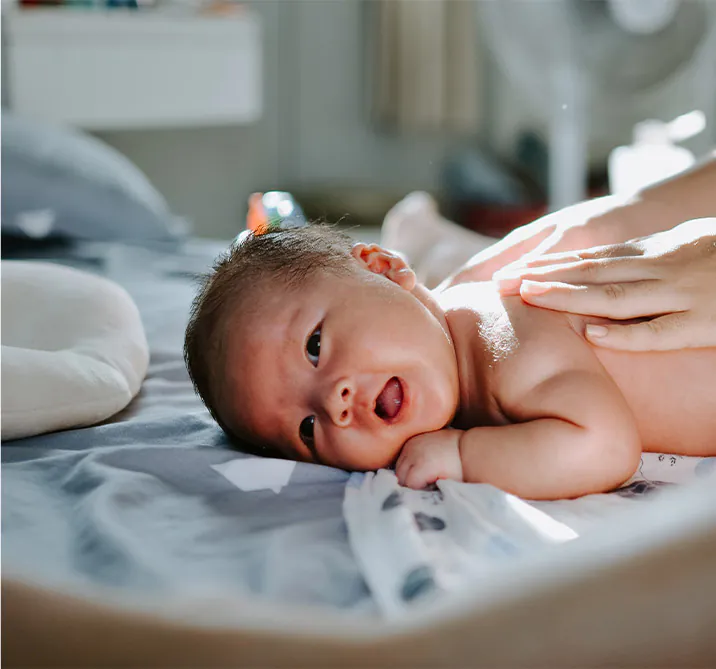A happy baby lies on a bed, smiling while being gently touched by a hand, with soft lighting in the background.