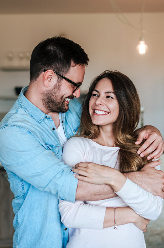 A happy couple embraces each other, smiling warmly, with a cozy indoor setting in the background.