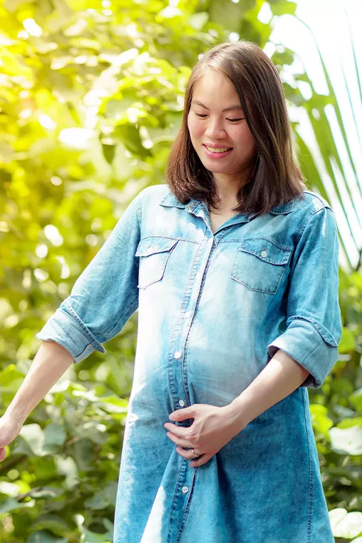 A smiling woman gently cradles her baby bump while surrounded by lush greenery.