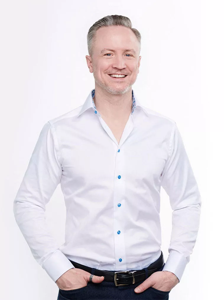 A smiling man in a white shirt with blue accents, standing confidently with hands on hips against a light background.