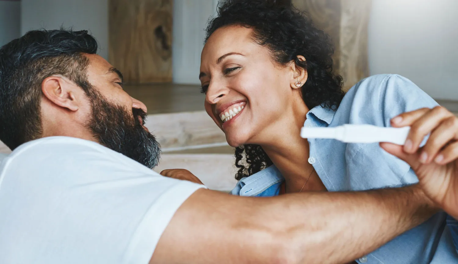 A joyful couple shares a lighthearted moment, smiling and looking at each other while one holds a pregnancy test.