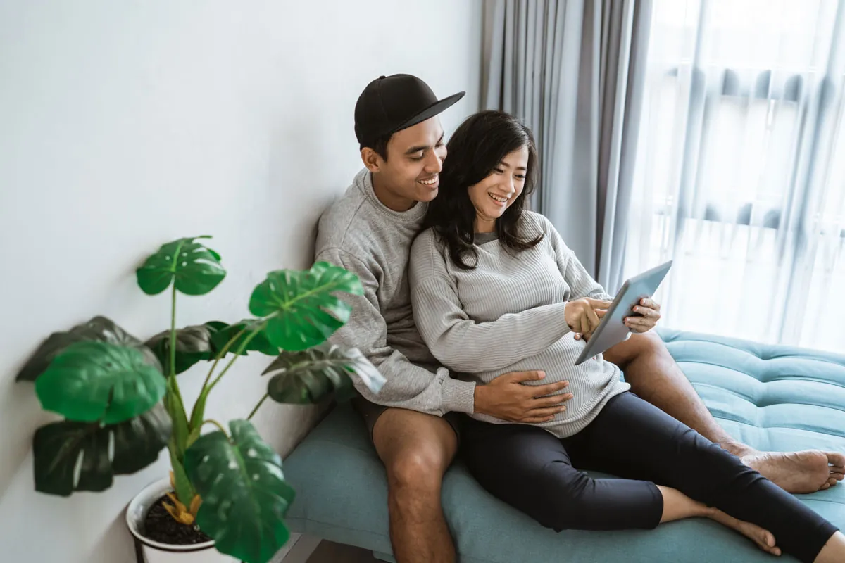 A couple sits on a blue couch, looking at a tablet together, with a green plant nearby and natural light coming through a window.