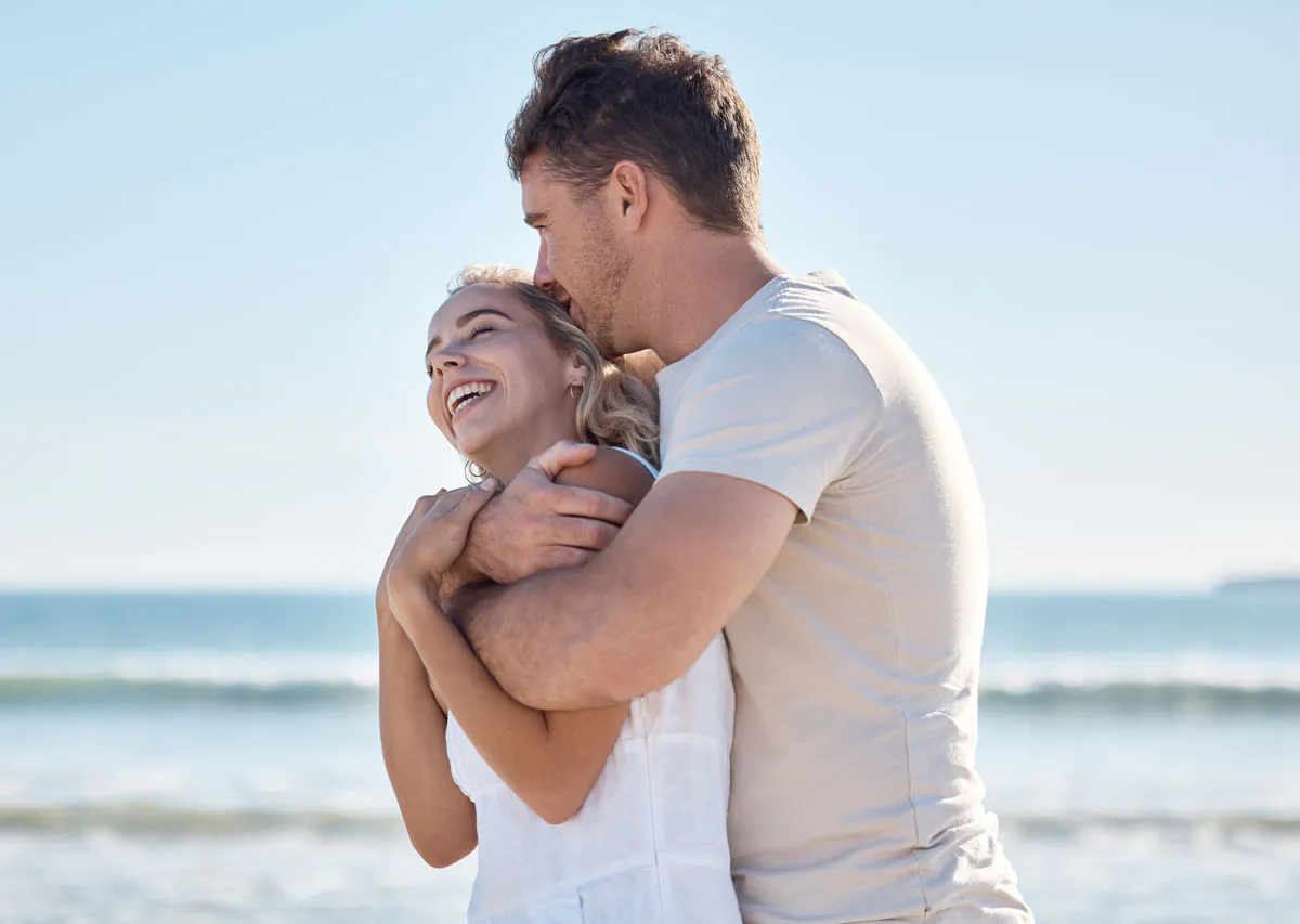 A joyful couple embraces at the beach, laughing and enjoying each other's company against a serene ocean backdrop.